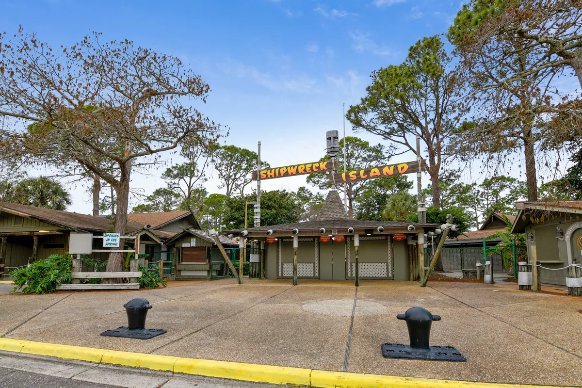 This image captures the entrance to Shipwreck Island Water Park, featuring the iconic sign, themed entrance structure, surrounding trees, and a welcoming paved pathway. The vibrant atmosphere is evident, suggesting excitement and family-friendly fun.