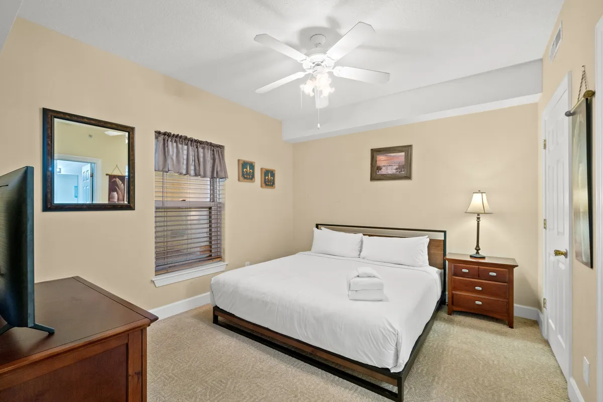 A tranquil bedroom featuring a queen-size bed with plush linens, a nightstand with a lamp, a wall-mounted flat screen television, framed artwork, and a window adorned with curtains. The space radiates comfort and offers ample natural light, creating an inviting retreat perfect for unwinding after a day of adventure.
