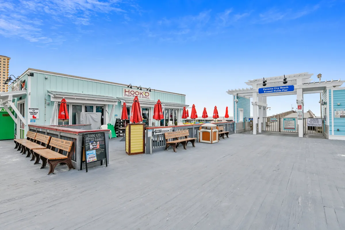 A lively boardwalk scene featuring the popular restaurant 'Hook'd' with red umbrellas, the entrance to the pier, and people dining outdoors. The casual, inviting ambiance invites groups to enjoy Panama City Beach’s dining and nightlife options.