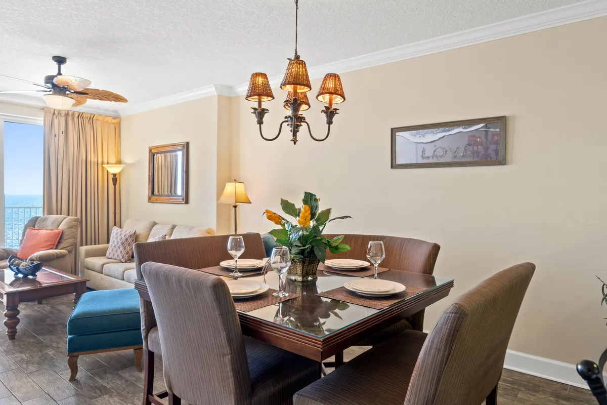 An inviting open-plan living and dining area with a stylish chandelier above the dining table, plush sofa, and expansive windows revealing an ocean view. The scene includes a cozy setup for group gatherings, with the dining table ready for meals and the living area ideal for casual relaxation or movie nights.