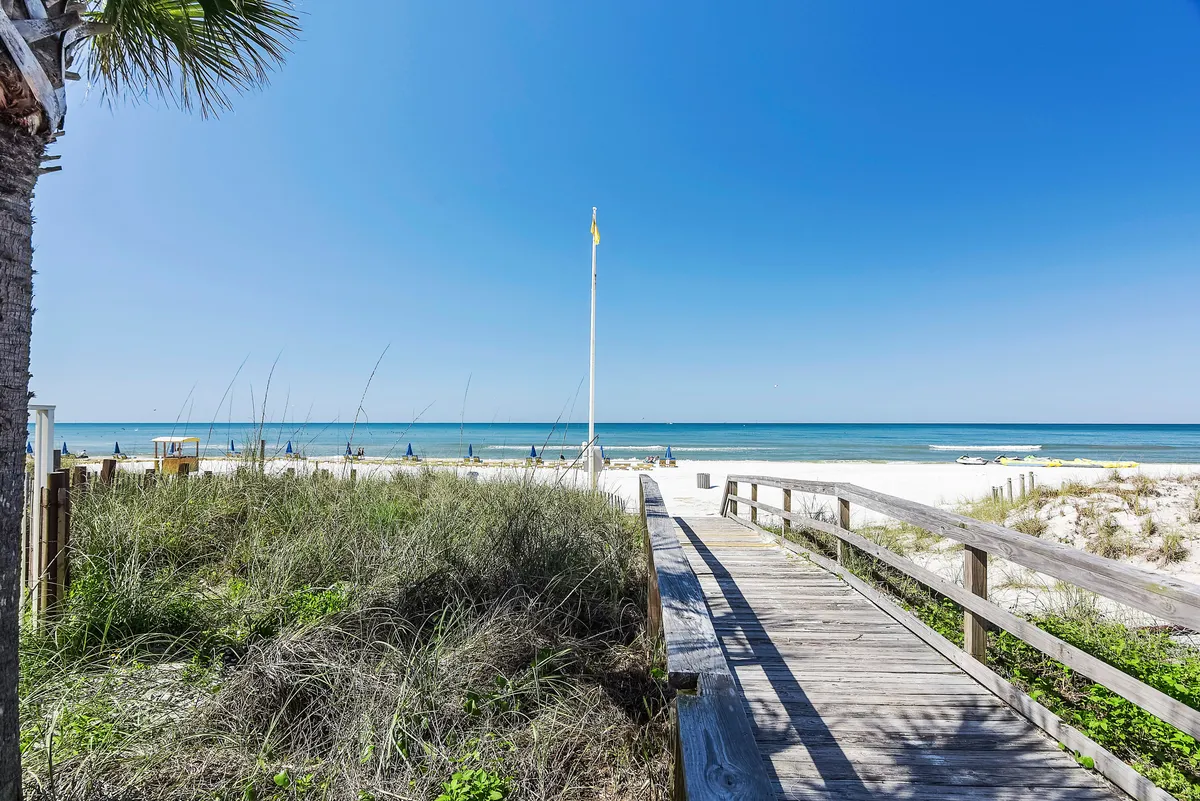 A scenic wooden boardwalk lined with palm trees leading to the beach, with umbrellas dotting the sand and the ocean stretching beyond. The setting is ideal for morning walks or sunset strolls, evoking a sense of peaceful exploration and access to nature.