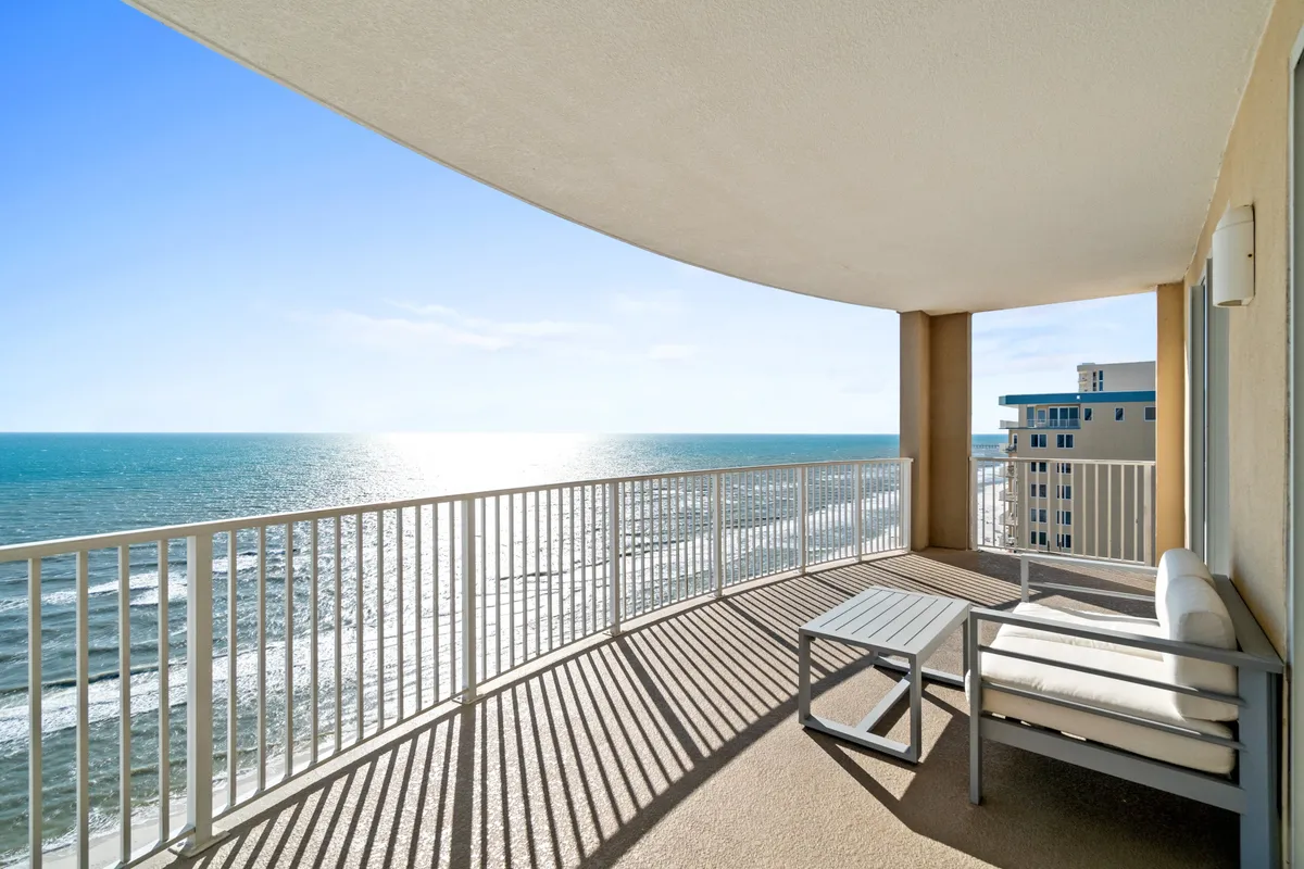 This image features a private balcony with a curved white railing, stylish outdoor seating with cushions, and shadows cast by the railing on the balcony floor. The panoramic ocean view dominates the background, with the blue horizon stretching as far as the eye can see. The setting feels serene and exclusive, inviting relaxation and contemplation.