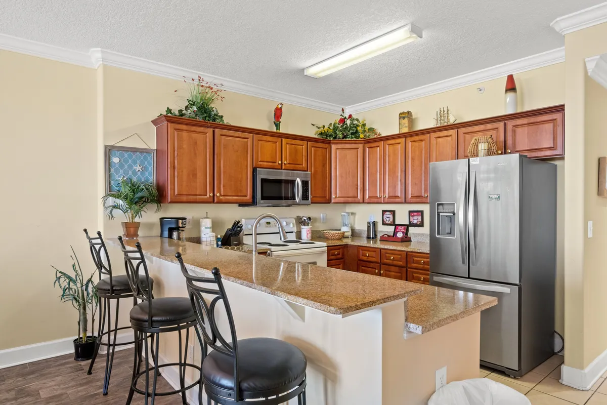 A granite breakfast bar with comfortable stools takes center stage in this kitchen, complemented by a stainless steel refrigerator, wooden cabinetry with decorative plants, and a microwave conveniently placed above the stove. The warm lighting and practical layout foster a sense of homey comfort and sociability.