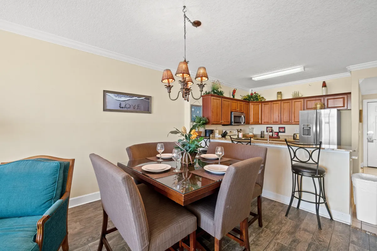 This image features a glass dining table elegantly set with plates and wine glasses, adjacent to a kitchen area boasting warm wooden cabinets and stainless steel appliances. A chandelier with soft, warm lighting hangs above the dining table, creating a welcoming and intimate setting. The arrangement suggests readiness for both casual breakfasts and special dinners.