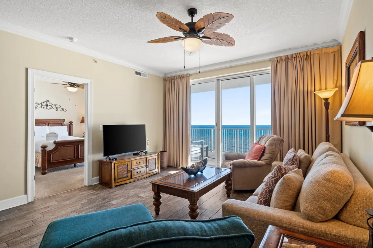 This image captures a spacious living room with a comfortable sofa and armchair, a wooden coffee table with a decorative centerpiece, and a television on a rustic stand. The room is filled with natural light from a large window that looks out to the balcony and features a stunning ocean view. The balcony doors invite the outdoors in, enhancing the sense of openness and connection to the coastline.