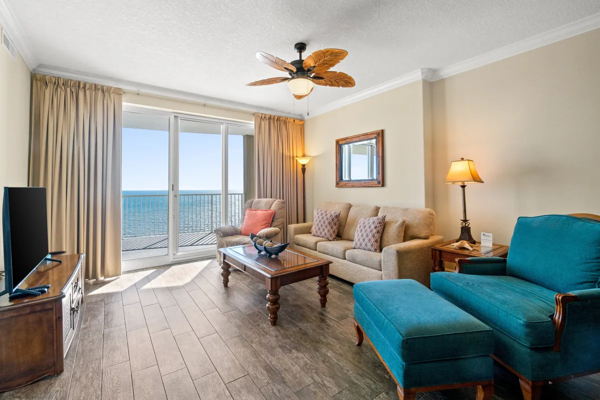 A bright, airy living space featuring large sliding glass doors opening onto an ocean-view balcony. The interior includes a blue armchair with ottoman, a beige sofa with patterned cushions, and a palm leaf ceiling fan, all arranged to maximize comfort while enjoying the view and natural light.