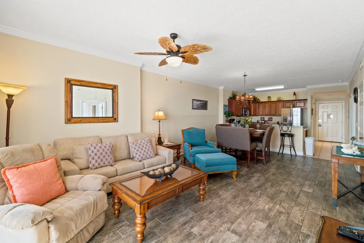 A plush beige sofa with patterned pillows and a turquoise armchair form a cozy living area, seamlessly connected to an open kitchen and dining space featuring bar stools. A wooden coffee table with a decorative centerpiece sits at the heart of the room, and the blend of soft furnishings and functional spaces creates a welcoming, stylish atmosphere.