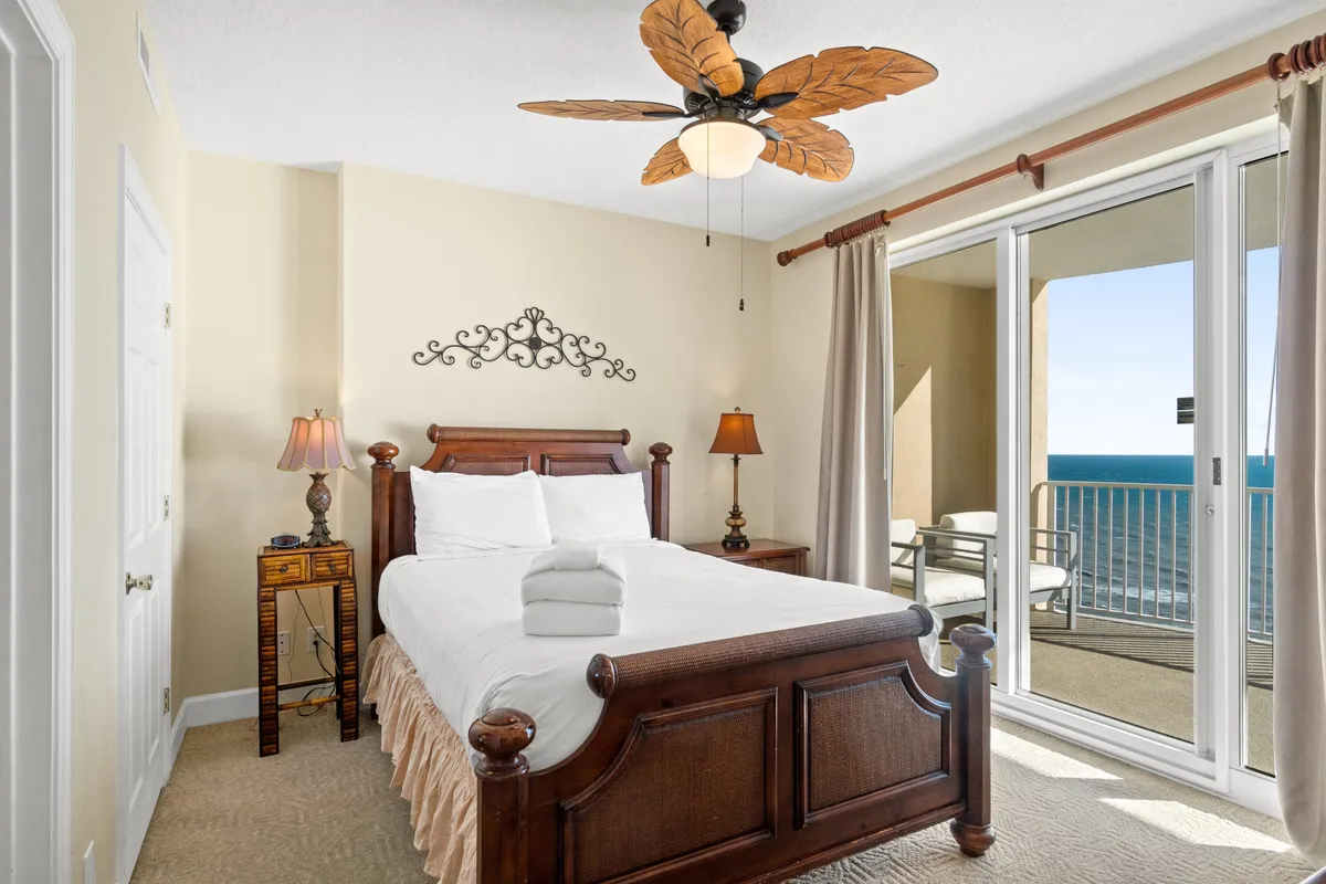 A stylish wooden bed with white linens and folded towels sits near a large sliding glass door, which opens to a private balcony with stunning ocean views. Ornate bedside lamps and a decorative ceiling fan create a tranquil and inviting atmosphere.