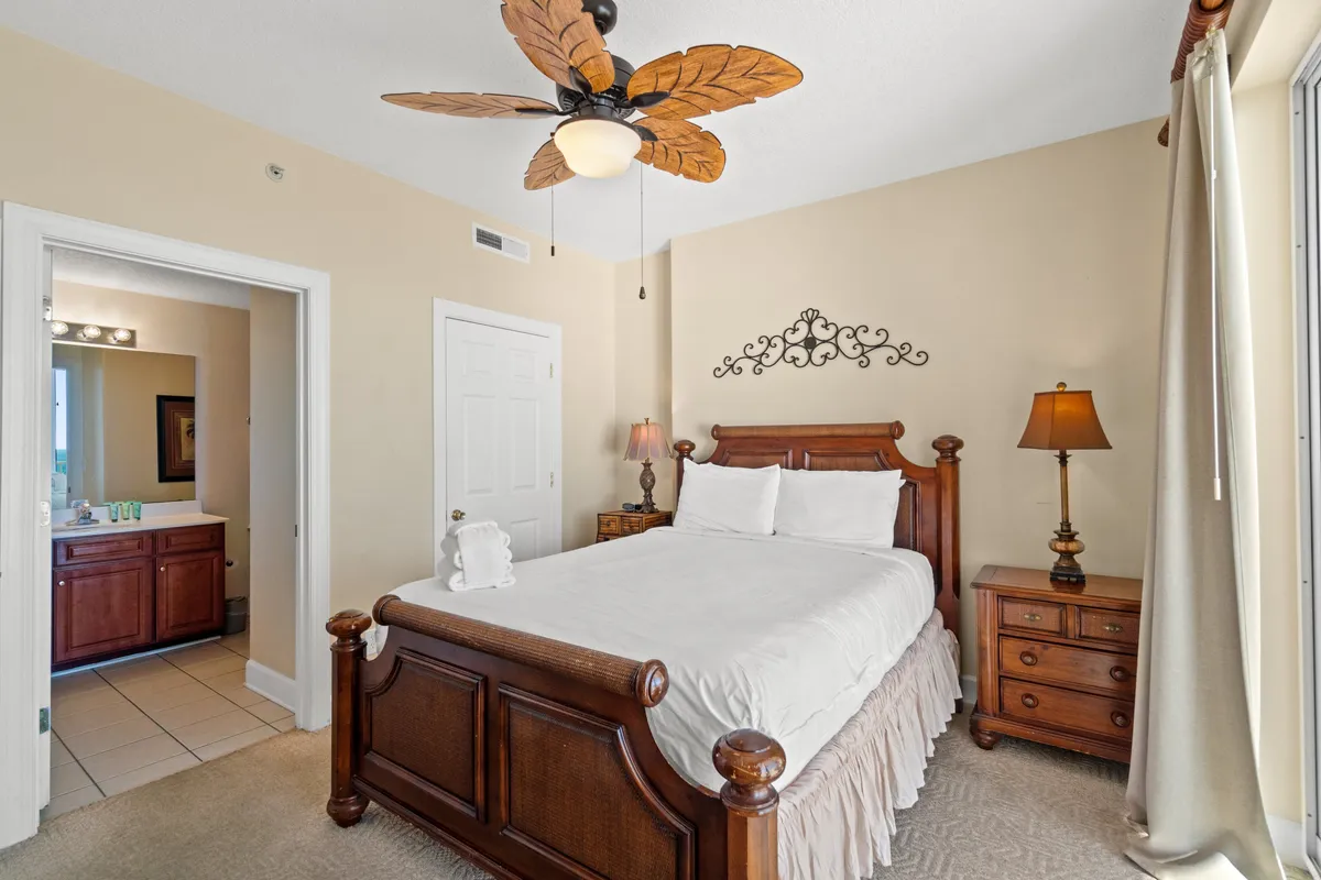A cozy bedroom with a wooden bed frame and white bedding, matching nightstands with lamps, decorative wall art, and a ceiling fan with leaf-shaped blades. The room feels inviting and restful, with soft lighting and natural textures.