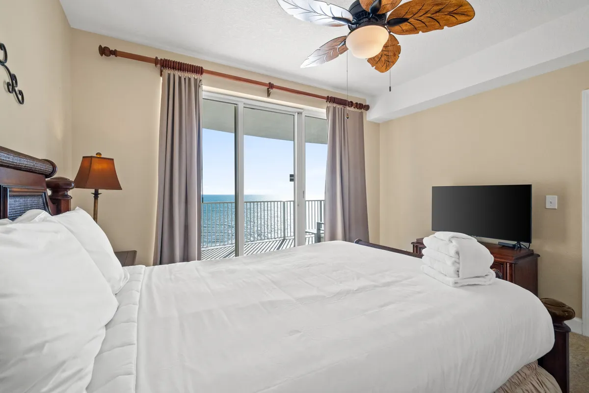 A large bed with crisp white linens faces sliding glass doors that open onto a balcony with direct ocean views. The room is bathed in natural light, and a ceiling fan with leaf-shaped blades adds to the serene coastal feel. The soothing color palette and uncluttered design evoke relaxation and comfort.
