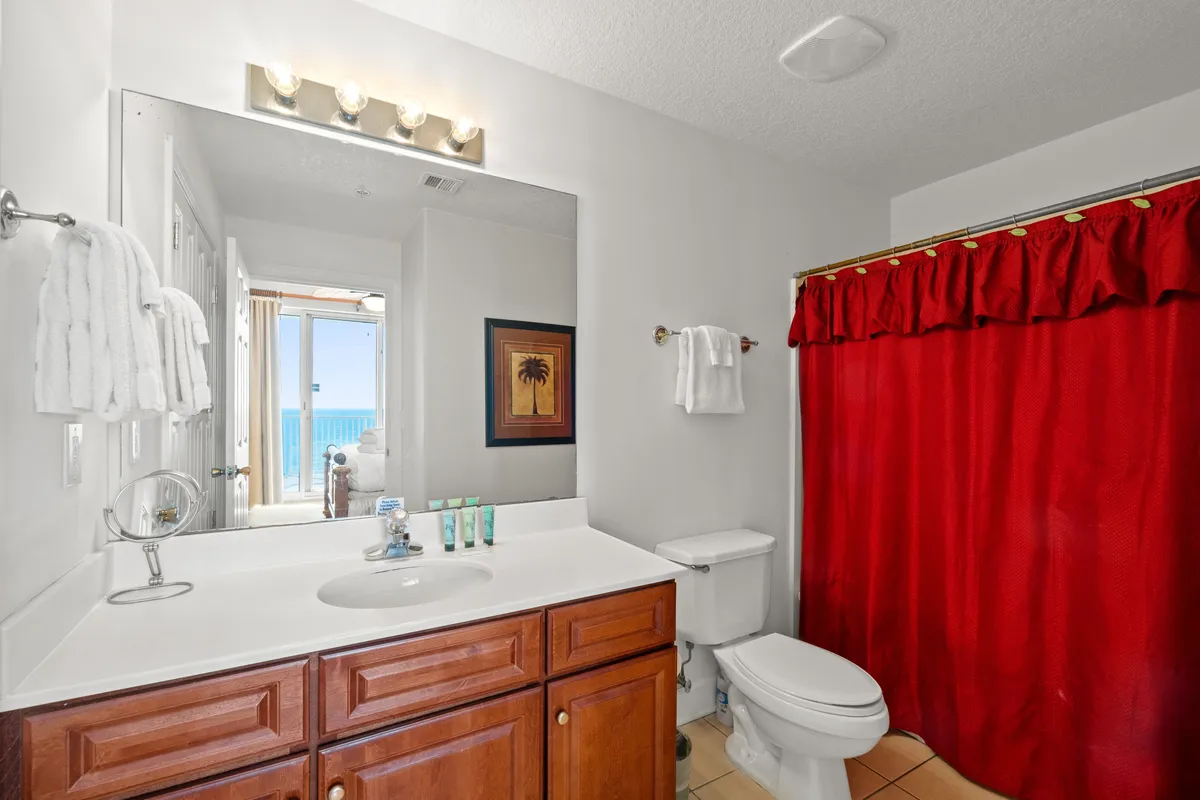A bright, fresh bathroom with a large mirror reflecting an ocean view, a vibrant red shower curtain, white fixtures, and wooden cabinetry. The room feels clean, practical, and welcoming with touches of coastal charm.
