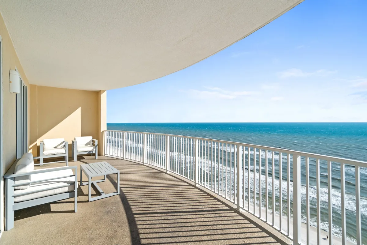This balcony features a curved railing casting unique shadows, cushioned outdoor chairs, and an expansive ocean view with rolling waves and a clear blue sky. The scene is peaceful, highlighting both the seating and the panoramic perspective.
