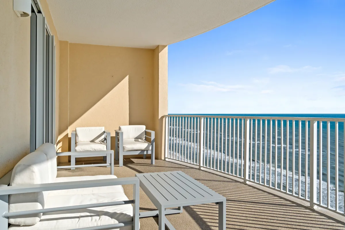 A private balcony seating area with white-cushioned chairs, a sofa, and a metal railing overlooks the ocean with visible waves. The setup suggests a perfect spot for lounging, reading, enjoying meals, or toasting a sunset. Shadows from the furniture and railings add to the relaxed, airy vibe.