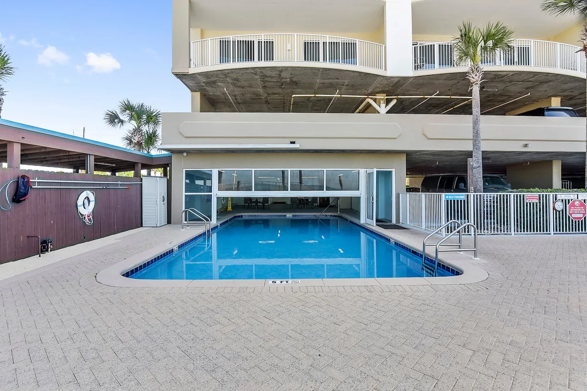 A rectangular swimming pool with clear blue water is partially covered by a retractable door, adjacent to a building with balconies and surrounded by palm trees. The setting suggests both indoor and outdoor enjoyment, with opportunities for swimming, lounging, and socializing in a tranquil resort environment.