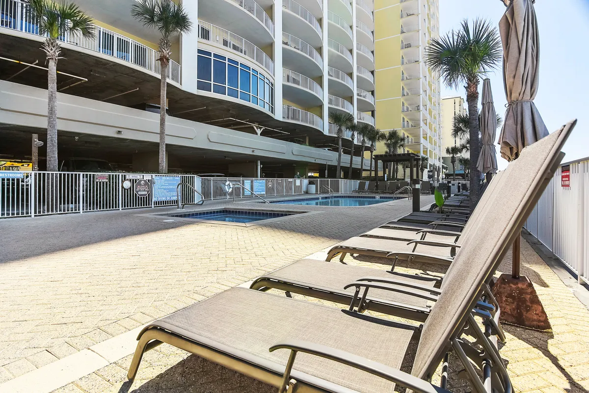 A sparkling swimming pool surrounded by lounge chairs with umbrellas, palm trees, and the towering resort building in the background. The inviting pool deck captures the essence of leisurely resort living and outdoor relaxation.