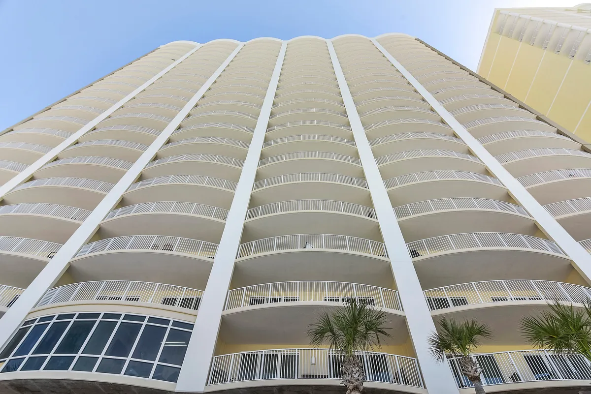 A high-rise condominium building stands tall by the beachfront, with rows of white railings on private balconies and palm trees at its base. The curved design of the balconies adds a sense of upscale, resort living and hints at the panoramic ocean views available from every unit.