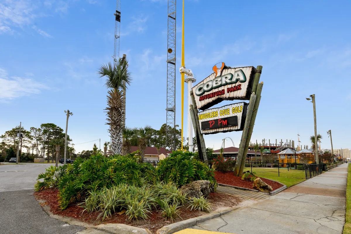 This image captures the vibrant entrance to Cobra Adventure Park, featuring its iconic sign, towering amusement rides, lush landscaping, and a welcoming sidewalk pathway. The scene is filled with energy, promising fun adventures and group activities.