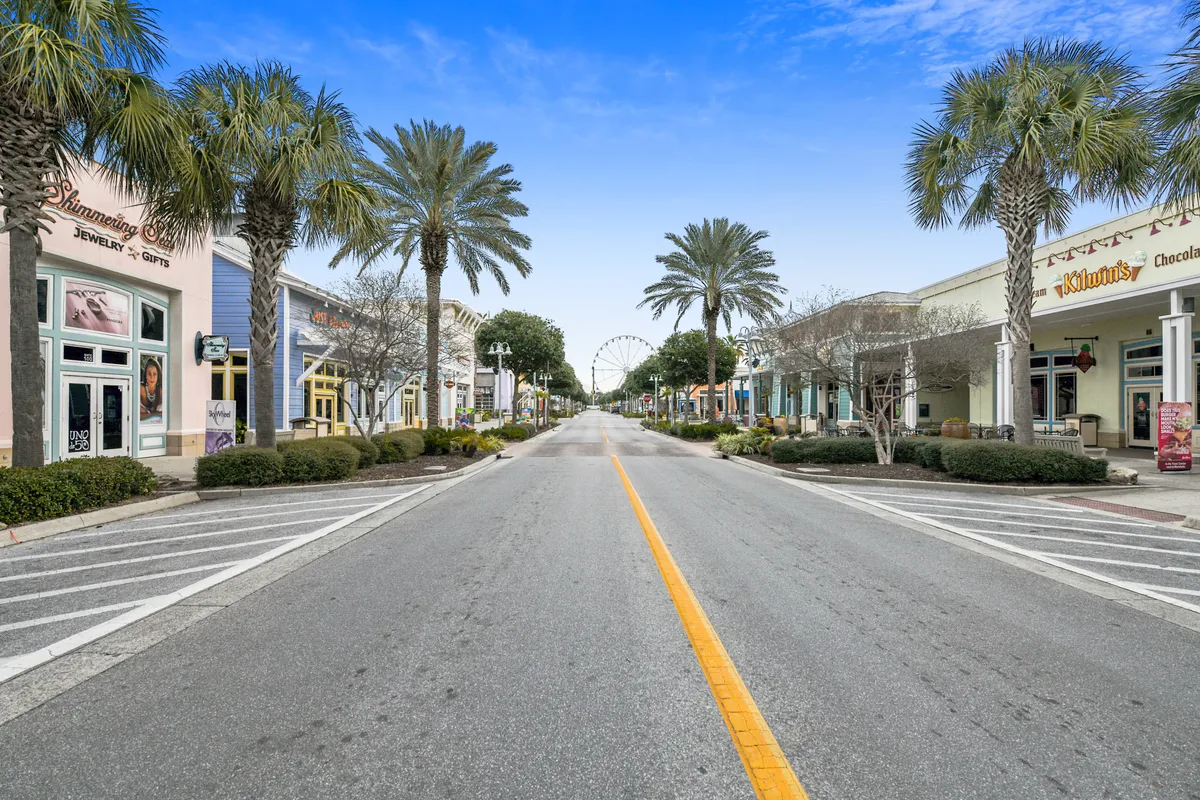 The photo showcases a wide, palm-lined street bustling with colorful storefronts and a distant Ferris wheel, set beneath a bright blue sky. The lively retail and entertainment district invites exploration, shopping, dining, and nightlife experiences with friends.