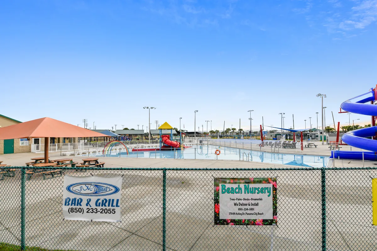 A vibrant, family-friendly outdoor area featuring a large pool with water slides, colorful playground equipment, and shaded picnic tables. The space is securely fenced, ensuring a safe environment for children to play and families to gather.